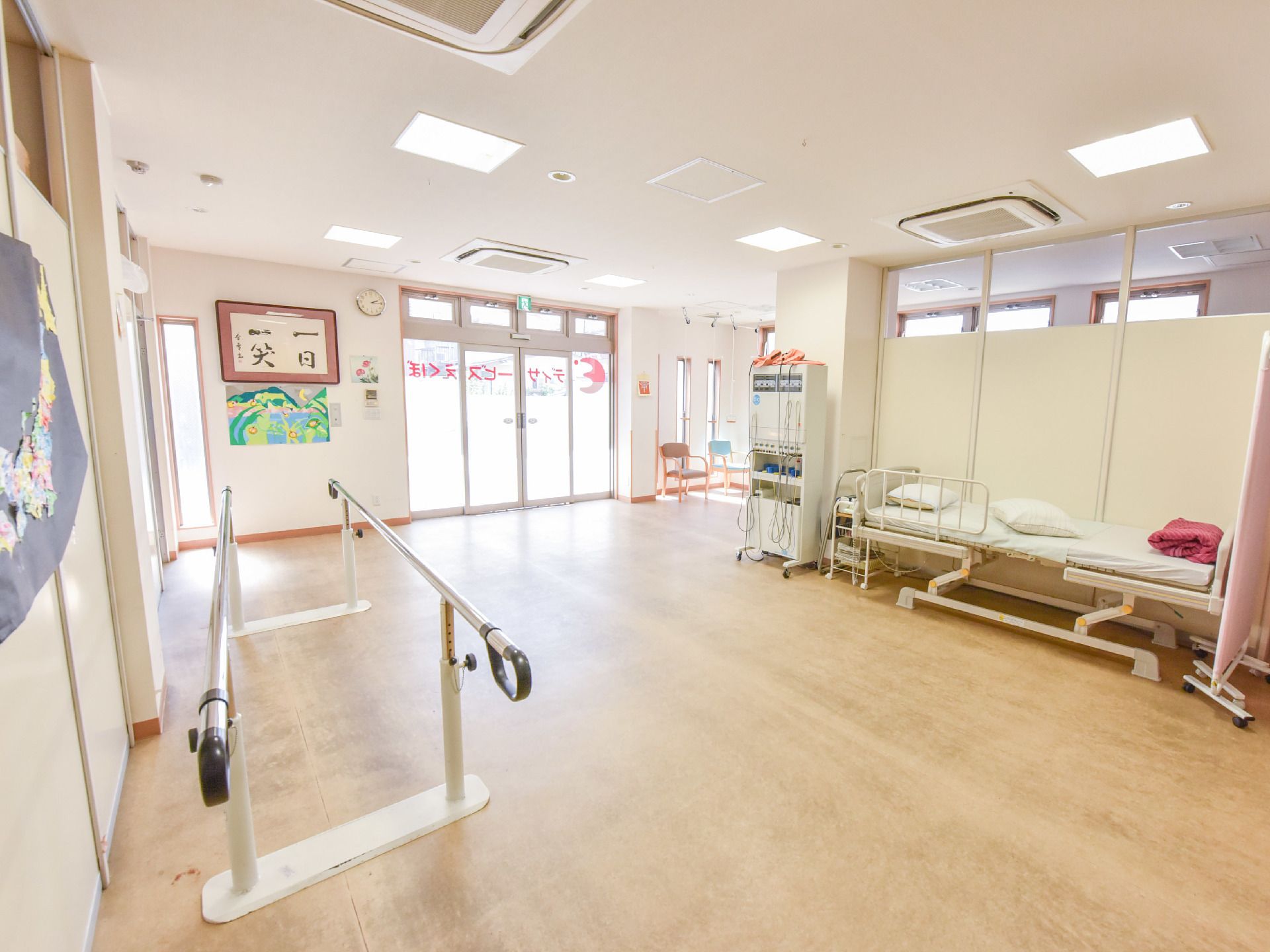 歩行用平行棒が置いてある。壁には、書が、飾ってある。ベッドがあり、落下防止の柵が取り付けてある。窓際に椅子が二脚見える。
