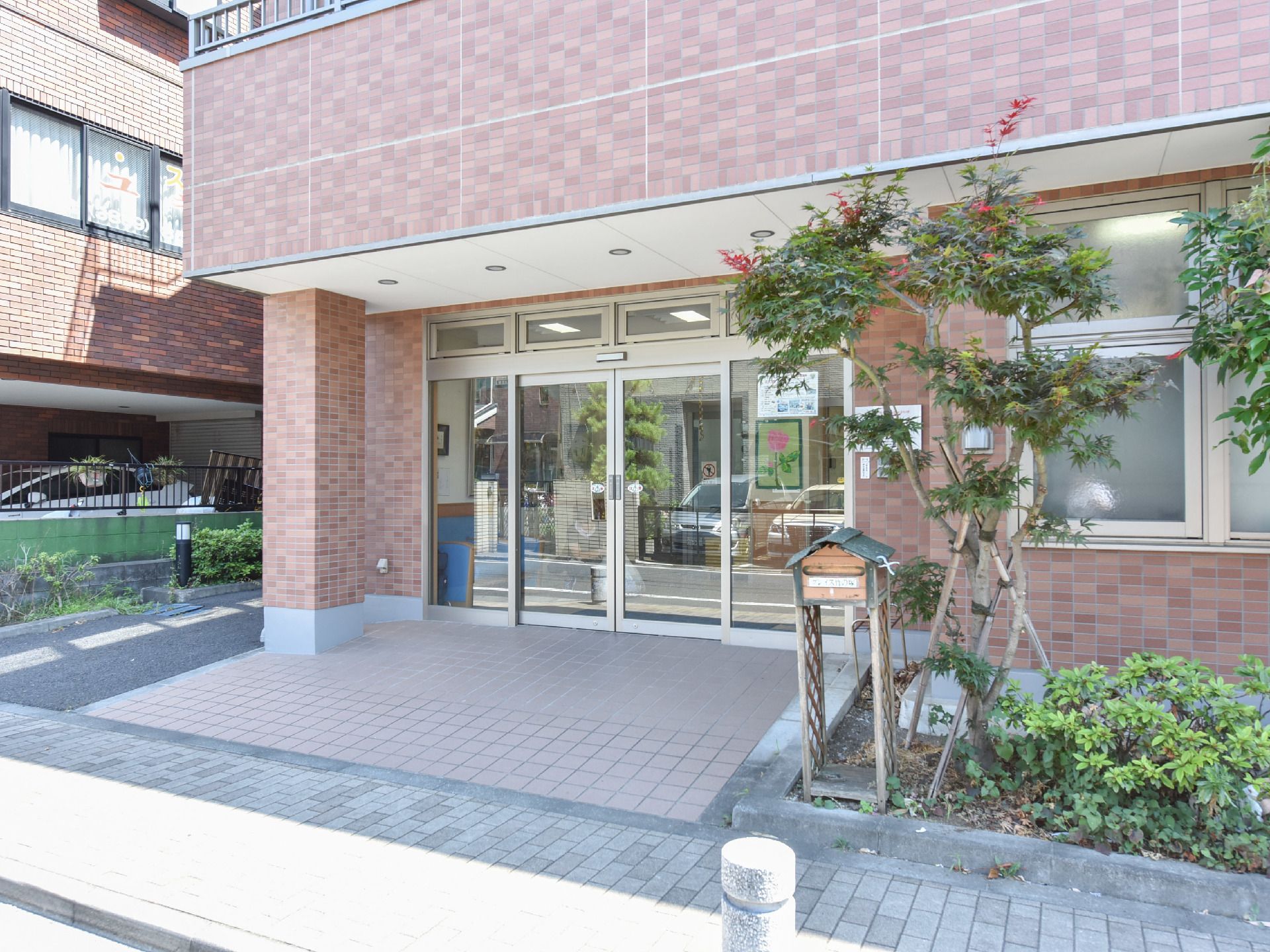 施設の外壁は茶色で、レンガのようになっている。黒いストライプのフェンスが隣の建物との間にある。花壇がある。