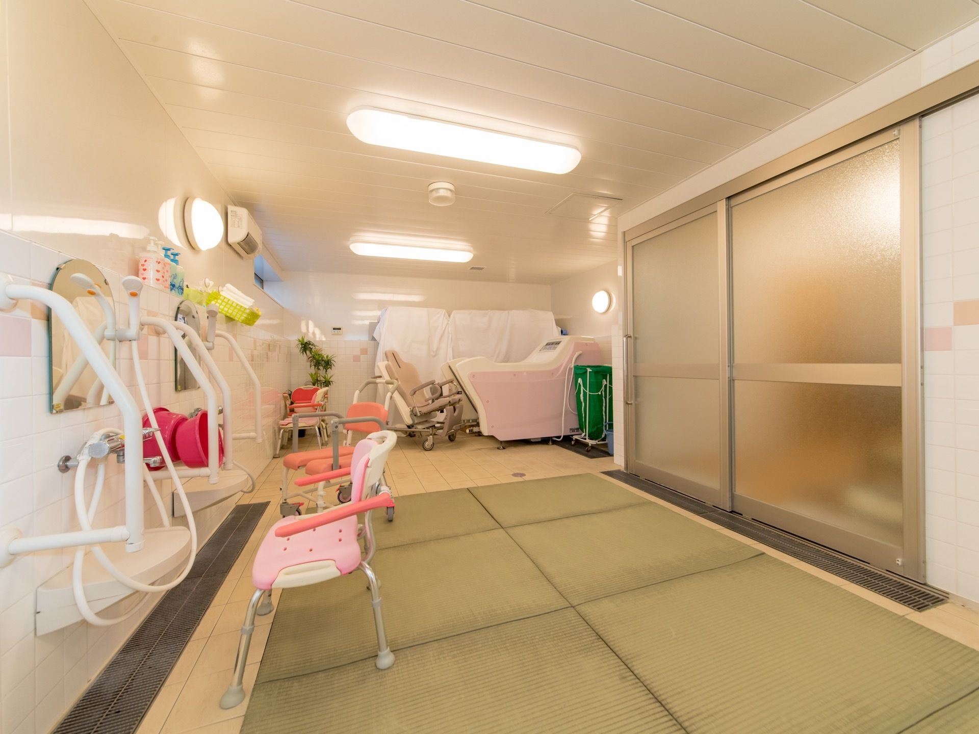 明るく広い浴室である。介護用の椅子とバスタブが置かれている。滑り止めのマットが敷かれている。各箇所に手すりが設けてある。