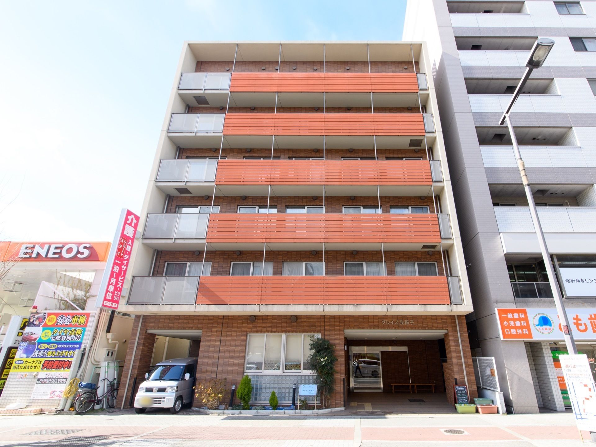 施設は、バルコニー付きの６階建て高齢者住宅である。１階部分に駐車スペースが設けられている。建物前には、プランターが置かれている。