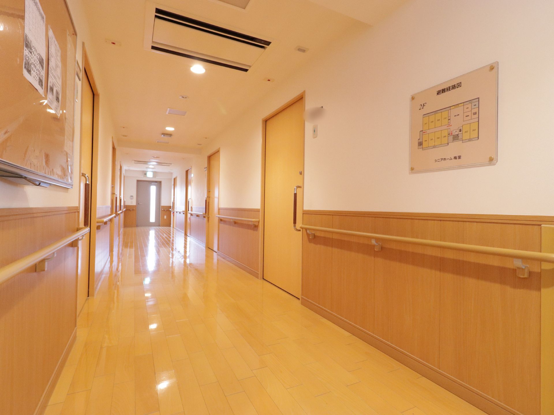 施設の廊下の様子である。壁には木製の手すりが取り付けられている。突き当りは直接外に出られる非常口となっている。
