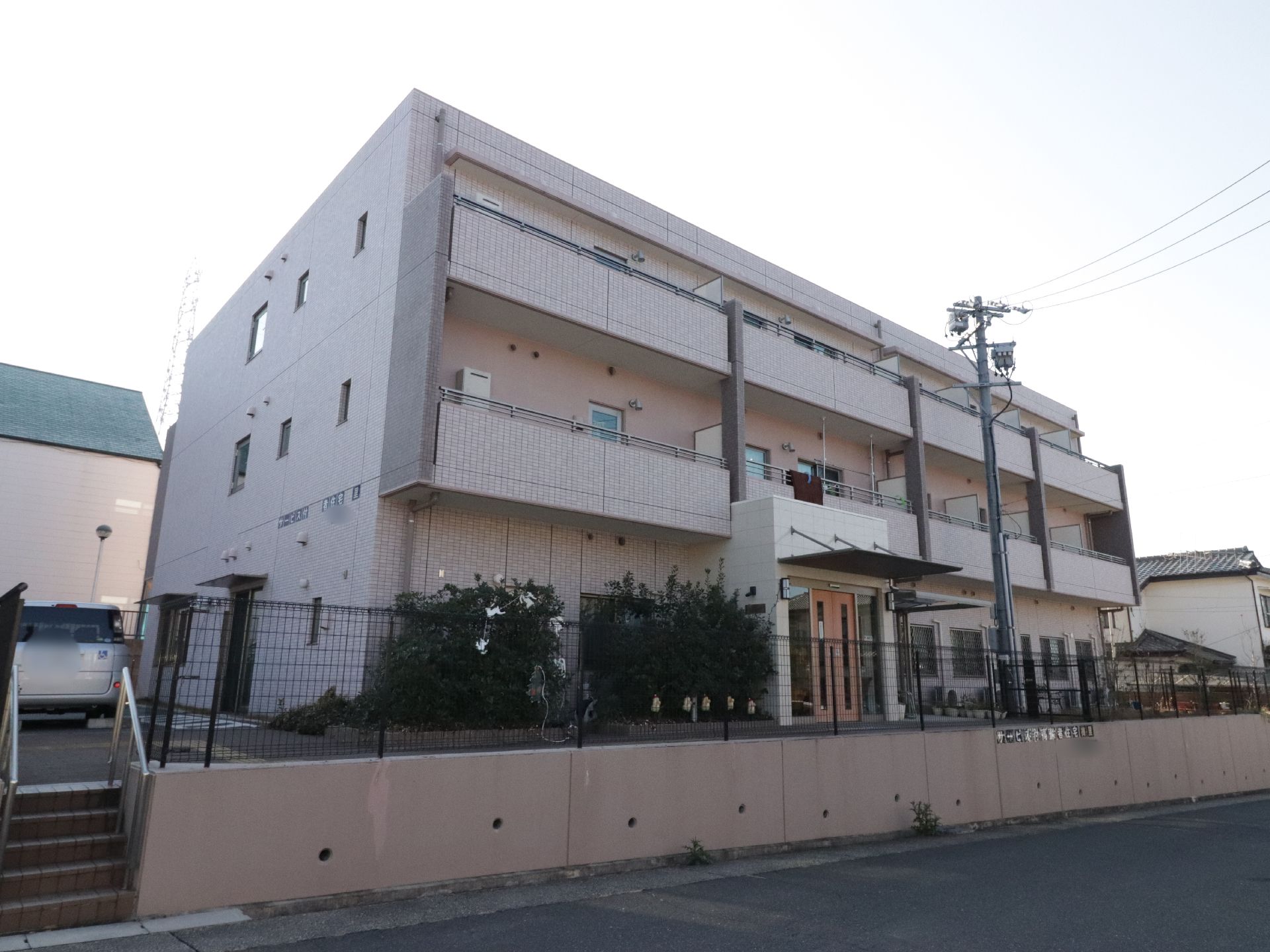 施設建物は鉄筋コンクリートの地上３階建て構造になっている。左側には駐車用のスペースが設けられ車が停まっている。