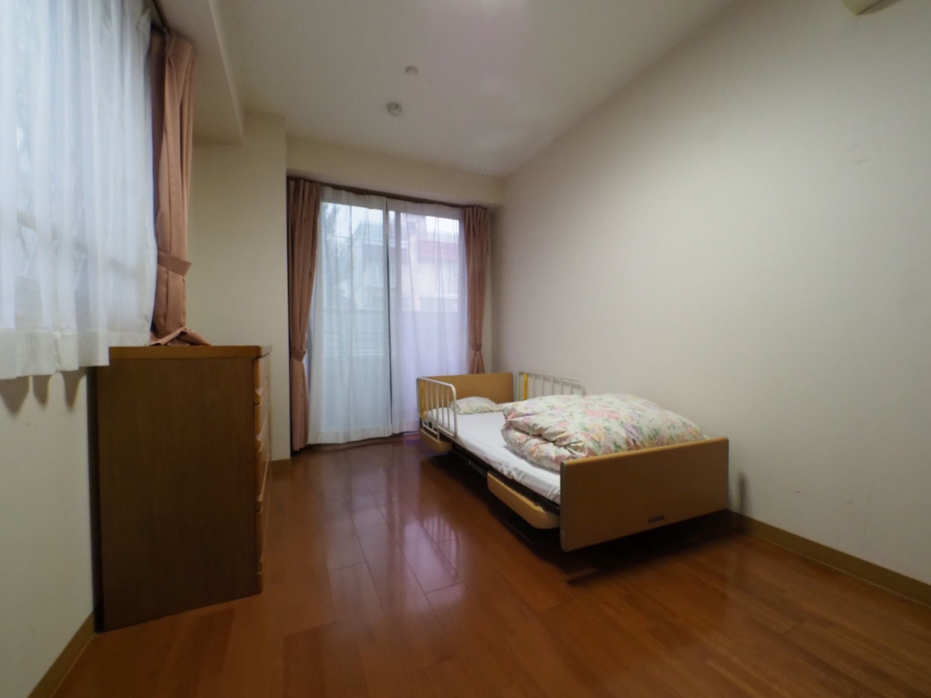 白い壁で窓があり、明るい居室になっている。窓にはピンク色のカーテンが掛けられている。床は落ち着いた色のフローリングになっている。