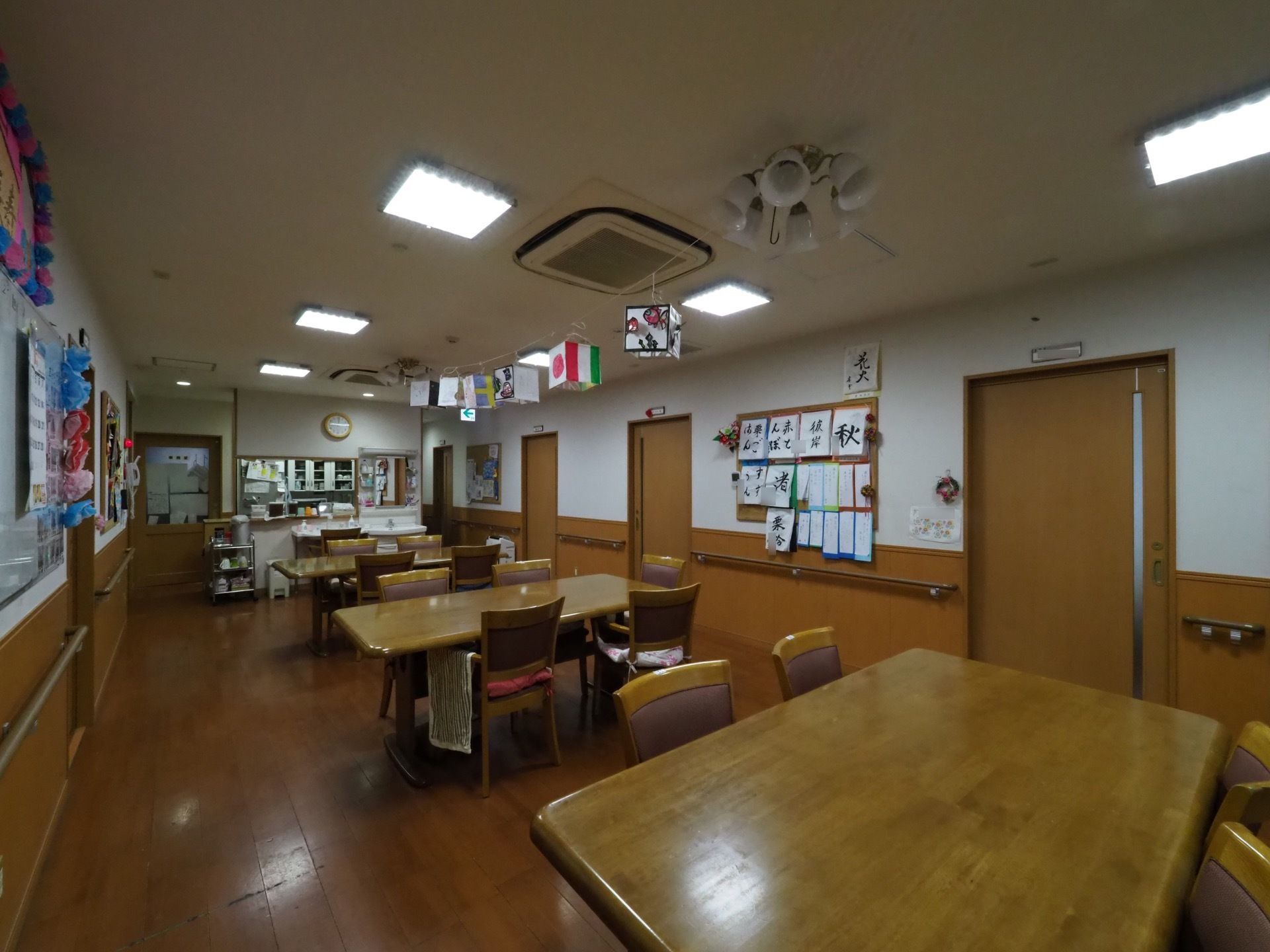 壁には入居者様の書道などの作品が飾られ、天井にも飾りがある明るい雰囲気のスペースになっている。テーブルと椅子がゆったり配置されている。