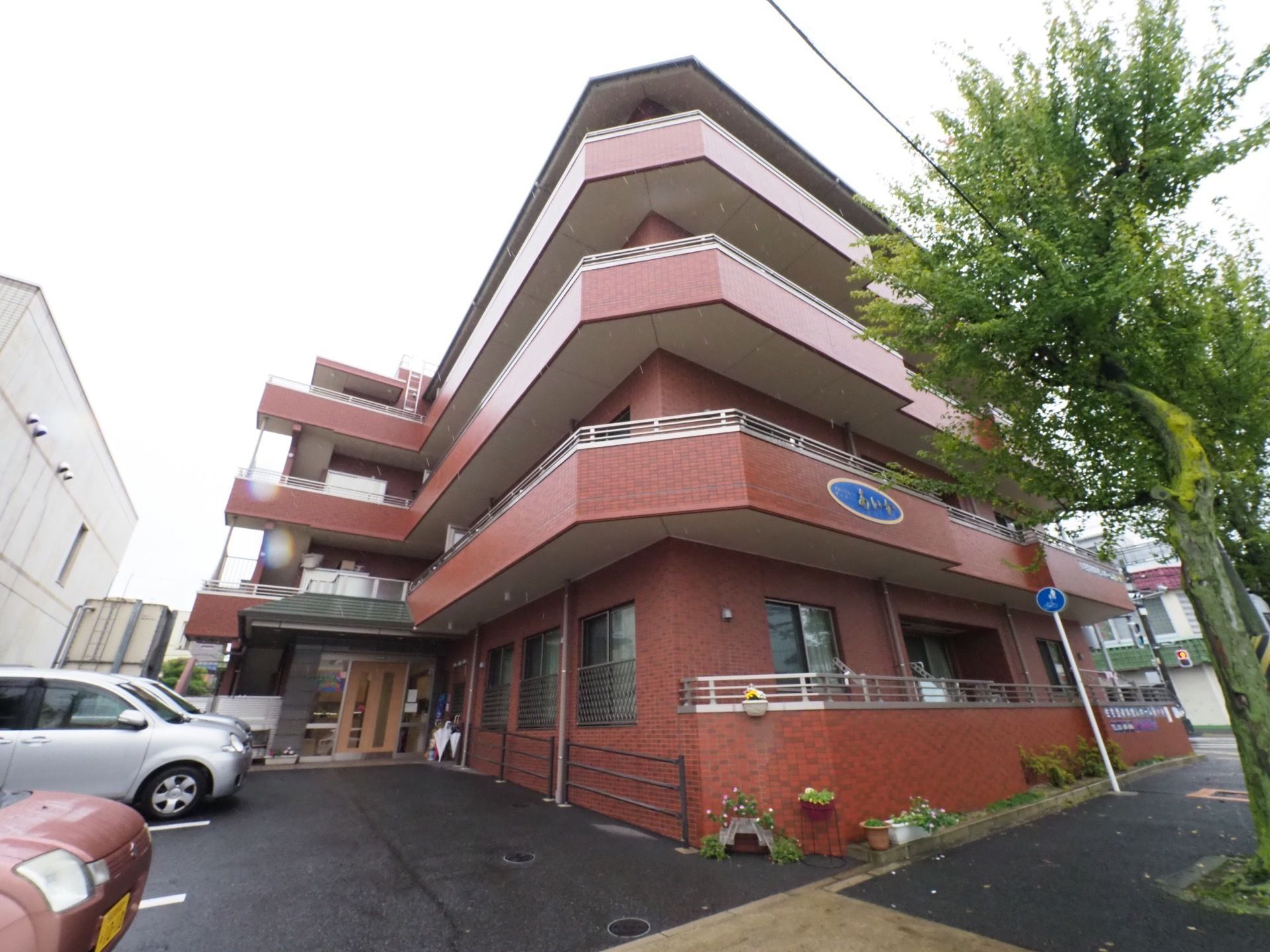茶色の外壁の建物で4階建てになっている。建物の前の歩道には大きな木があり、施設側には鉢植えなどが置かれている。