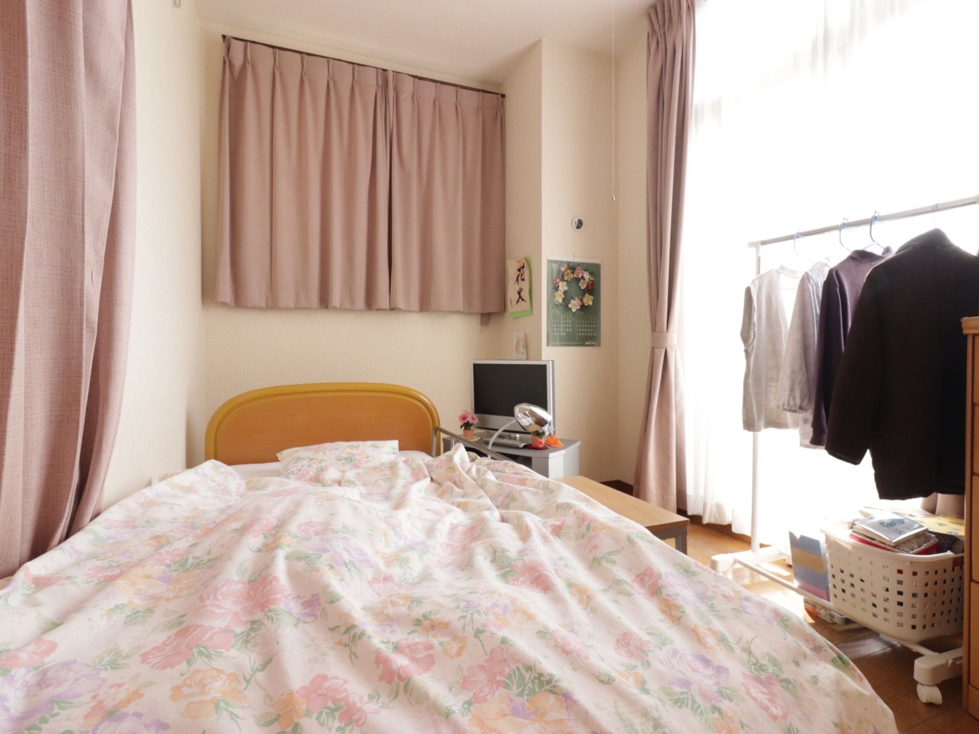 室内に、電動介護ベッドやテレビ、小さなテーブル、ハンガーラックが置かれている。大きな窓が付いており、明るい。