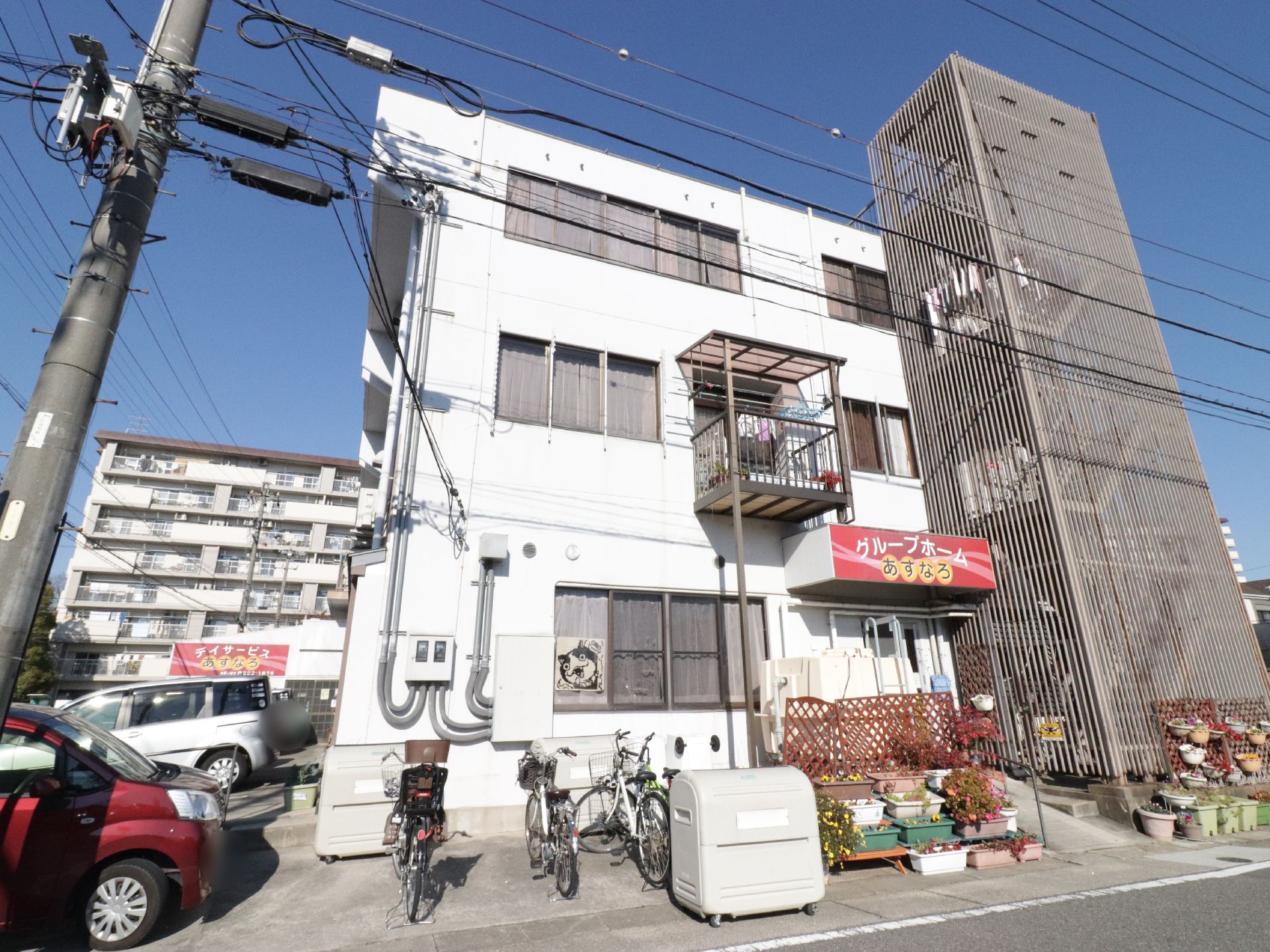 施設外観は道路に面して建てられていて、3階建てである。すぐ隣には車が数台止められている。部屋の窓にはカーテンが掛かっている。