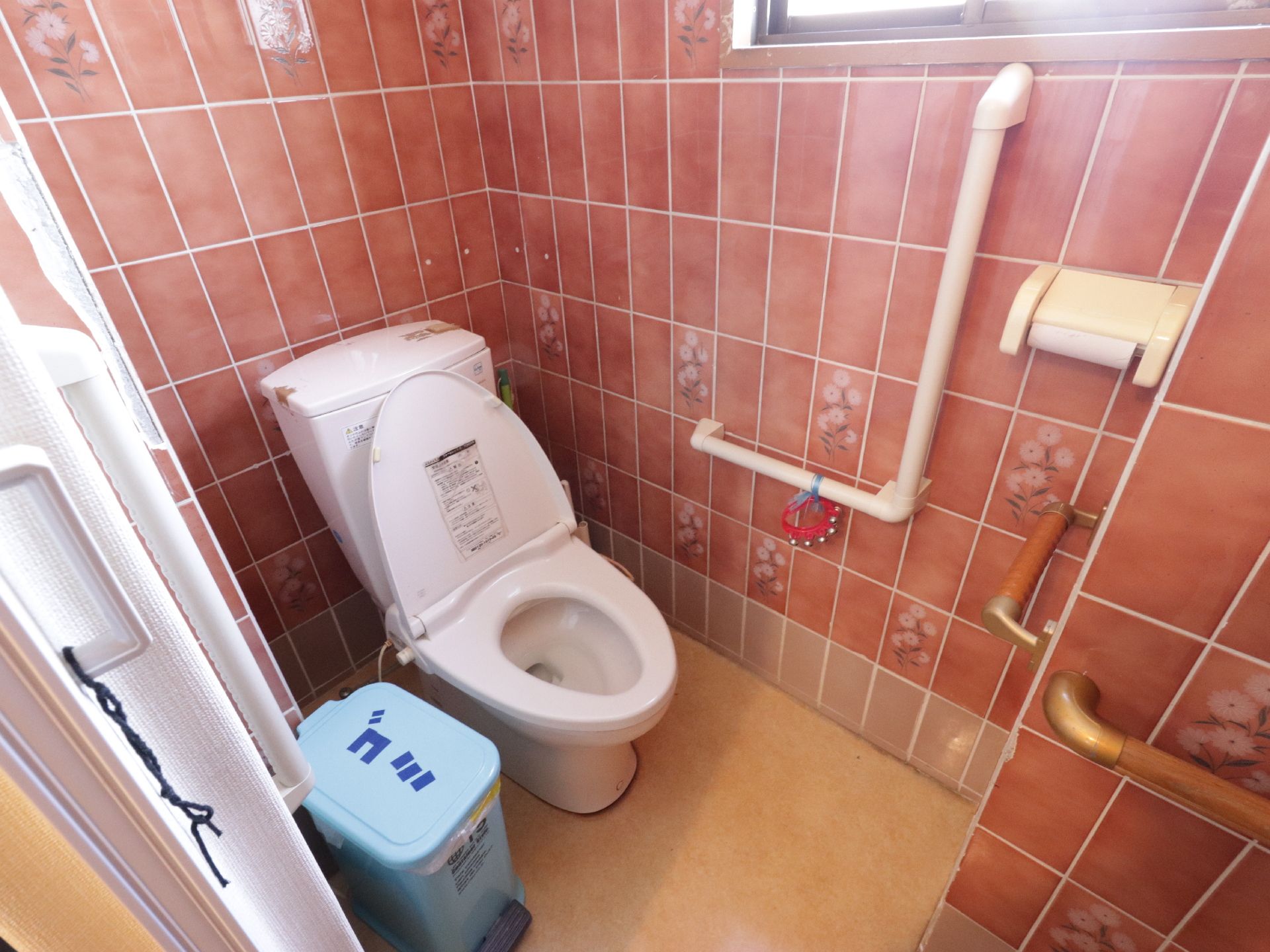 トイレには洋式便器が設置されている。L字型の手すりには鈴が取り付けられている。ゴミと書かれたゴミ箱がある。