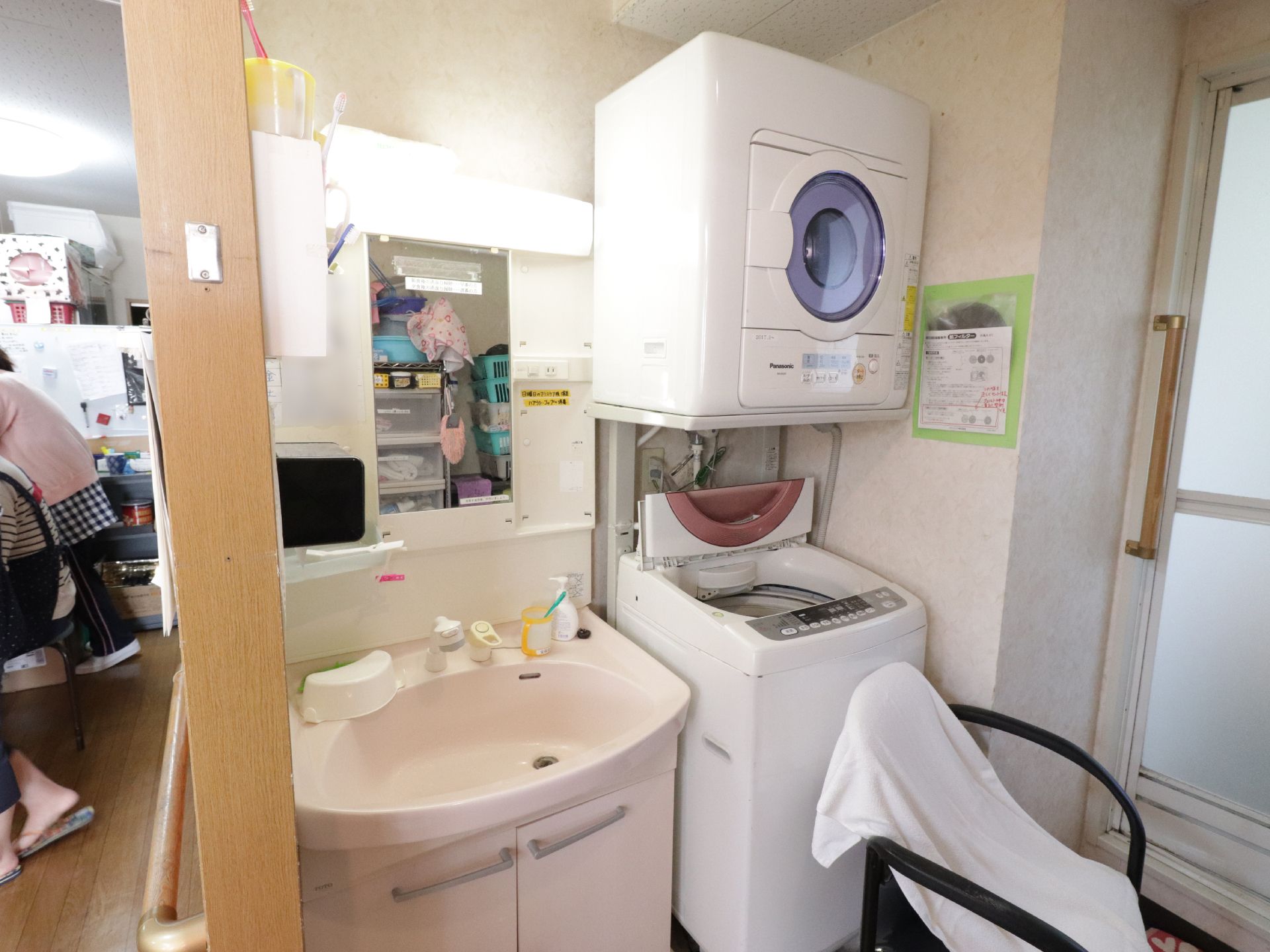 鏡付きの洗面台がある。その隣には洗濯機があり、その上には乾燥機が置かれている。背もたれ、ひじ掛け付きの椅子が置かれている。