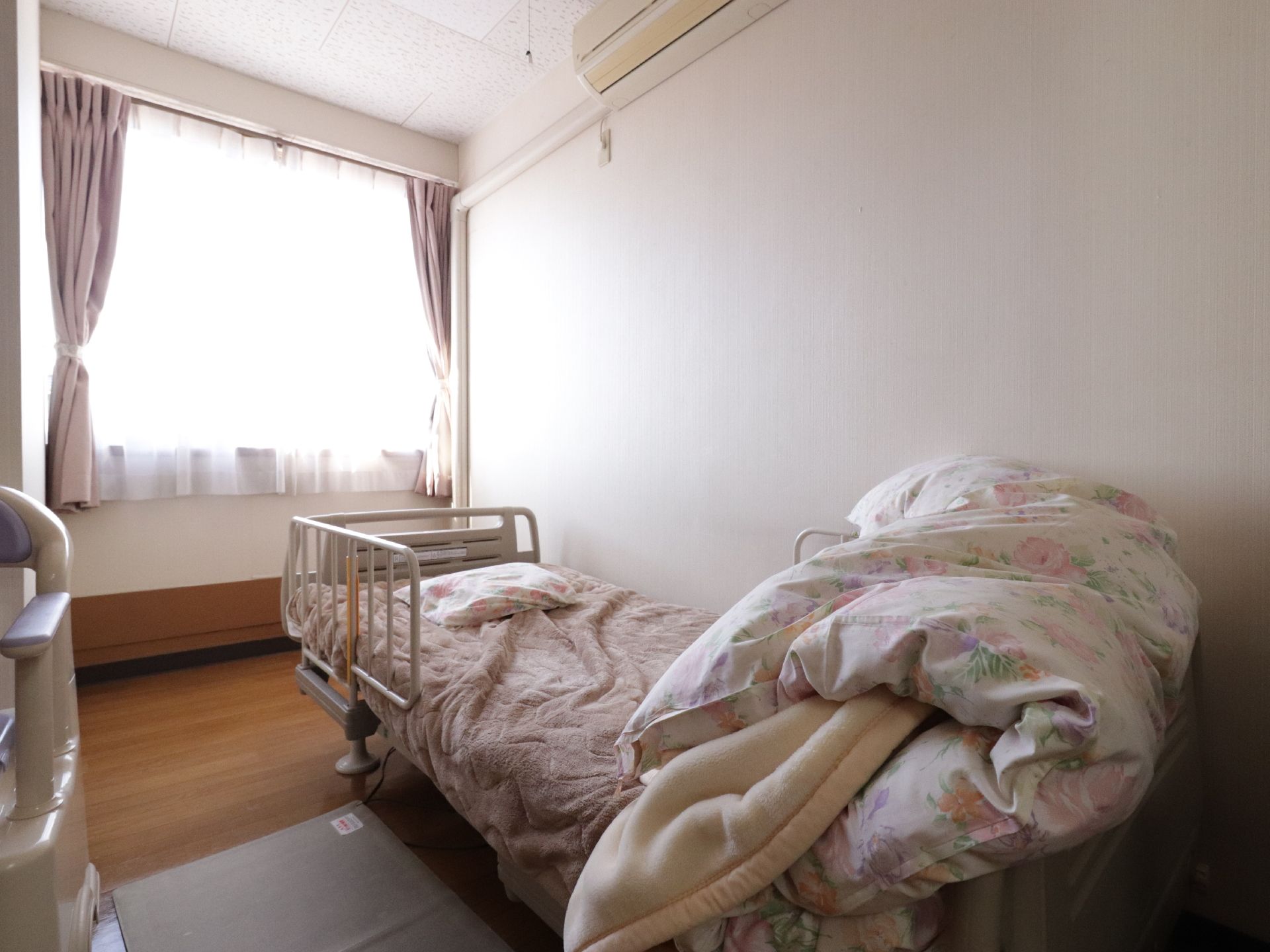 居室にある窓からは日が差し込んでいる。ベッドの布団は半分に畳まれている。ベッドの上の方にはエアコンが設置されている。