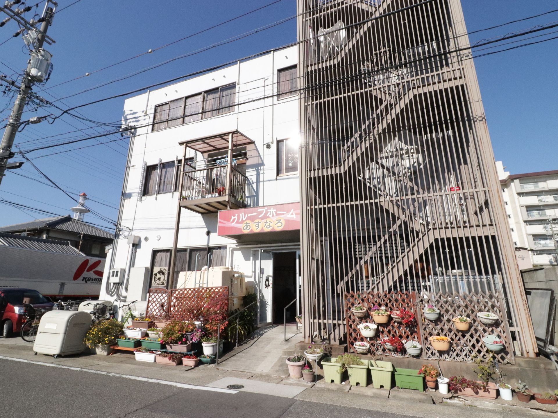 施設は道路に面して建てられている。施設名が書かれた赤い屋根がある。施設前にはプランターや植木鉢が複数並べられている。