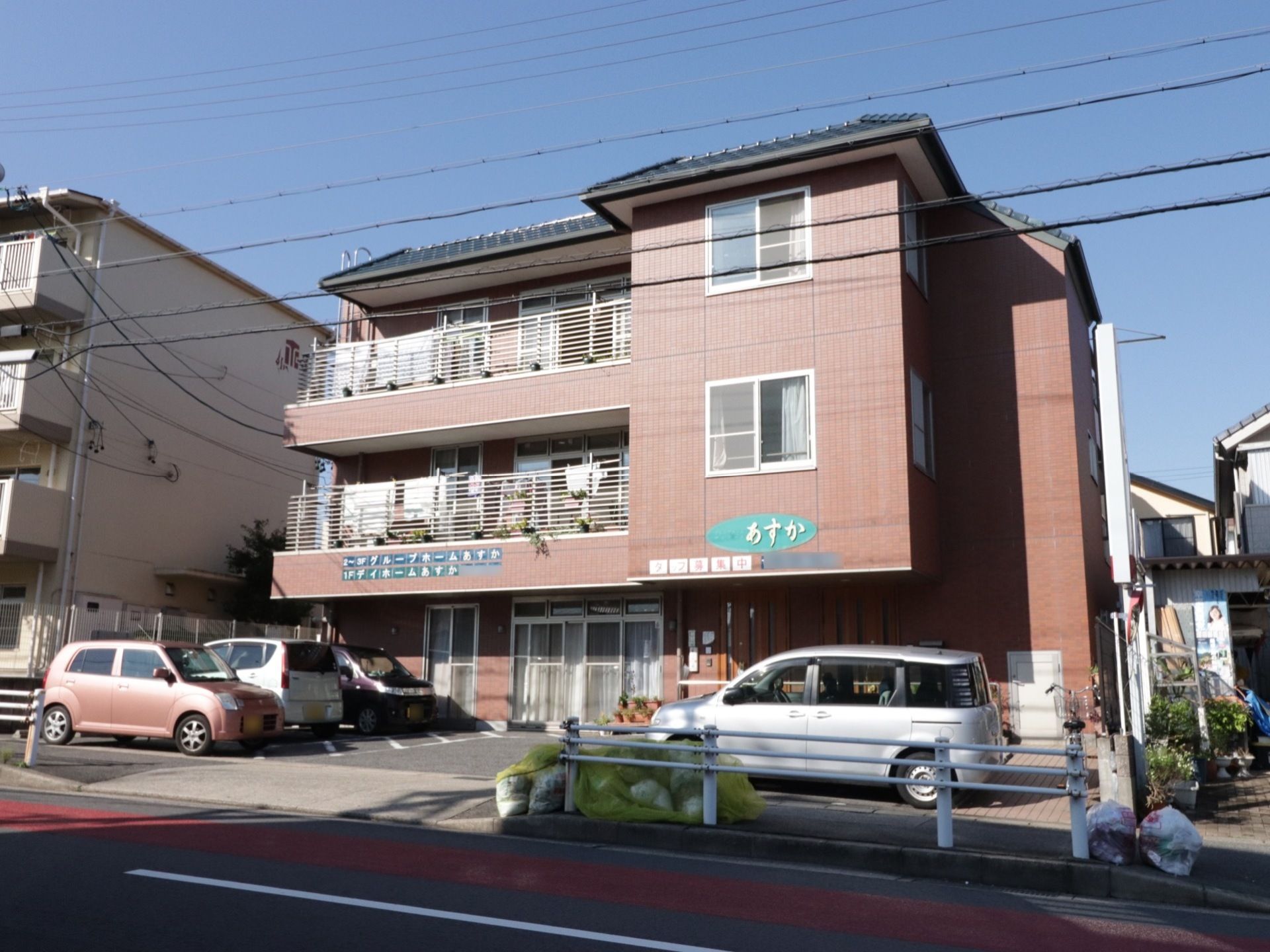 施設名が外壁にわかりやすく表記されている。建物の前に駐車場がある。周辺には高い建物がなく日当たりがいい。