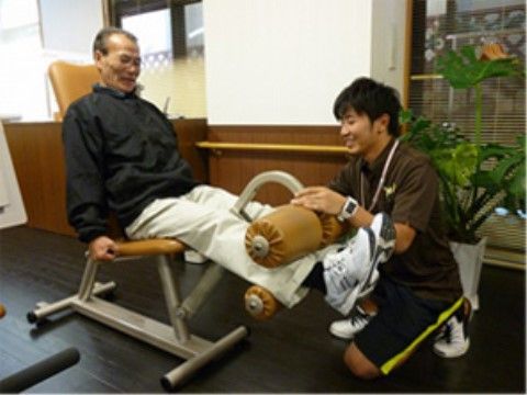 フィットネス器具を用いて足のリハビリに取り組む高齢男性と介助する男性職員