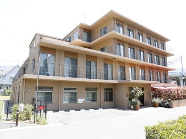 建物は4階建てである。外壁はベージュ色でテラスの手すりはグレー色である。建物前に3台、駐車スペースがある。玄関サイドに観葉植物がある。