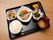 黒いお盆に並んでいます。左前から時計回りにご飯、小鉢、サバの煮つけインゲン、ニンジン添え、菜ものあえもの、くだもの、味噌汁。