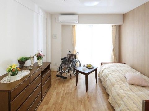 木を基調とした明るく落ち着いた雰囲気の居室である。備え付けられたベッドの幅は広い。ほか、収納用の棚やエアコンなどが設置されている。
