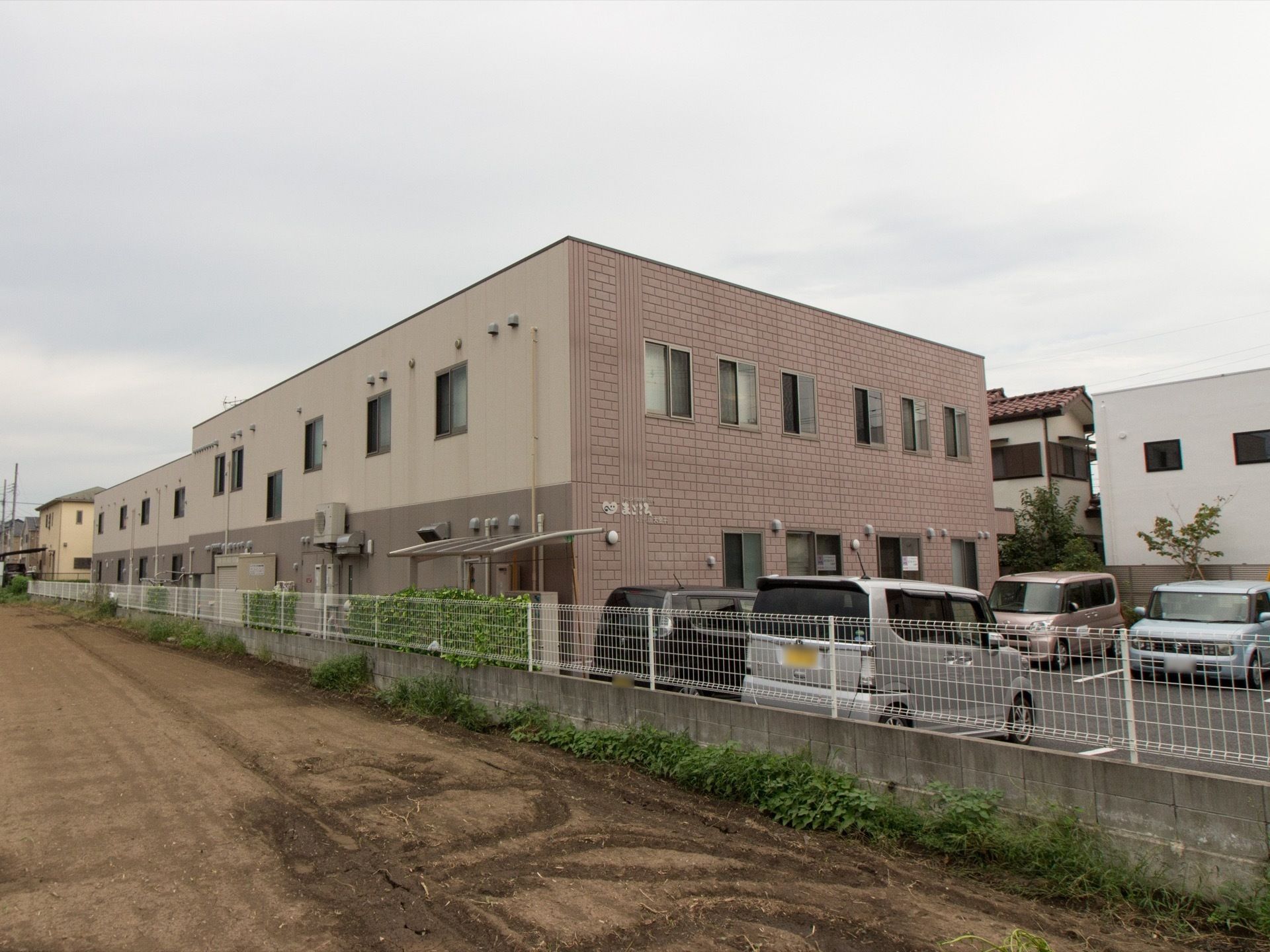 落ち着いたブラウン調の２階建て施設である。白いフェンスが設置されている。車が複数台停められる、駐車場も完備。