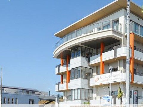 白い壁の大きな建物である。最上階のベランダは透明なフェンスが付き、半円形に飛び出している。ベランダ下と建物の端にはオレンジ色の柱がある。