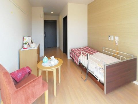 バリアフリーの行き届いた居室である。木目調の床と壁、白の壁紙で統一された落ち着いた印象。ベッド横にはナースコールもある。
