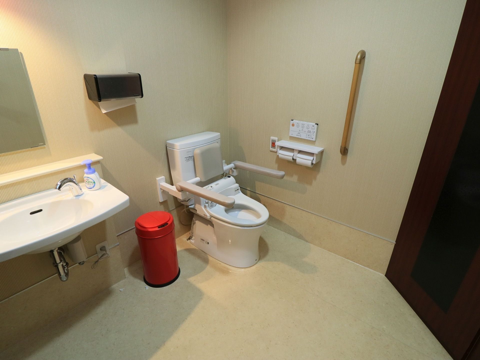 広いスペースを確保した共用トイレである。洗面台や手すり、背面付きアームレストが完備されている。呼び出しボタンや温水洗浄便座も設置されている。
