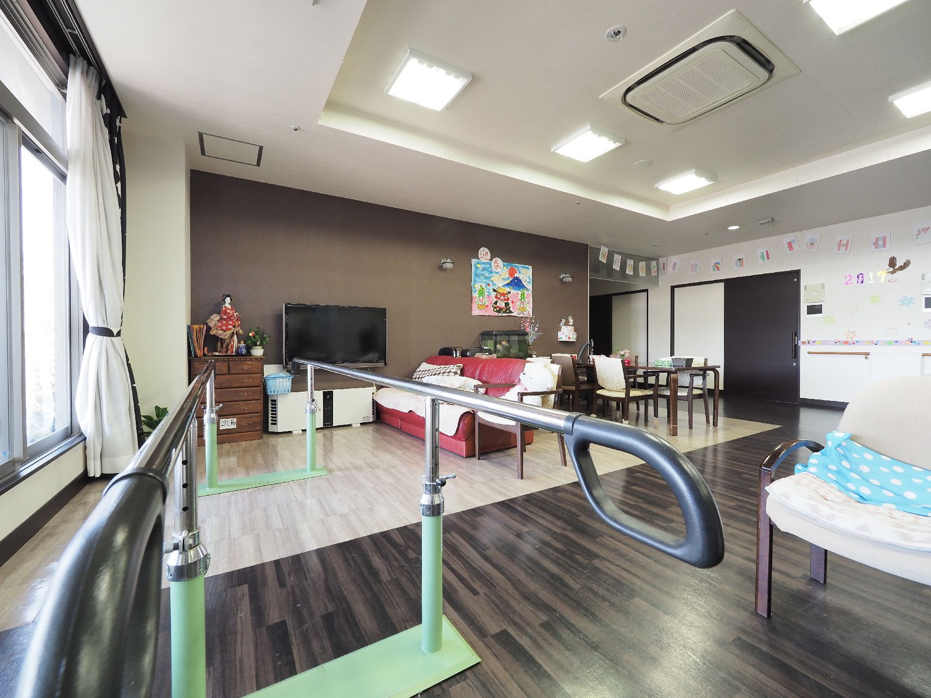 フロアの奥に、大型のテレビとソファ、その傍にダイニングテーブルが置かれている。大きな窓の近くには歩行訓練用の平行棒が設置されている。