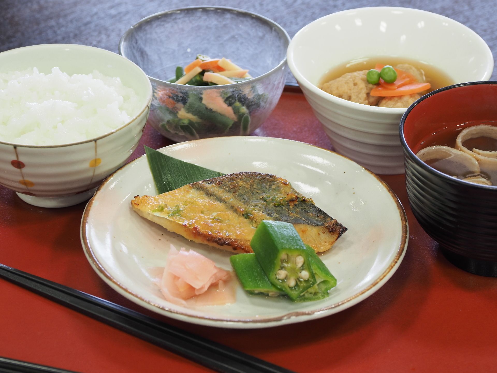 朱塗りのお盆にご飯と味噌汁、焼き魚の他に煮物と和え物の小鉢が置かれている。栄養バランスと彩りが考慮されたメニュー。