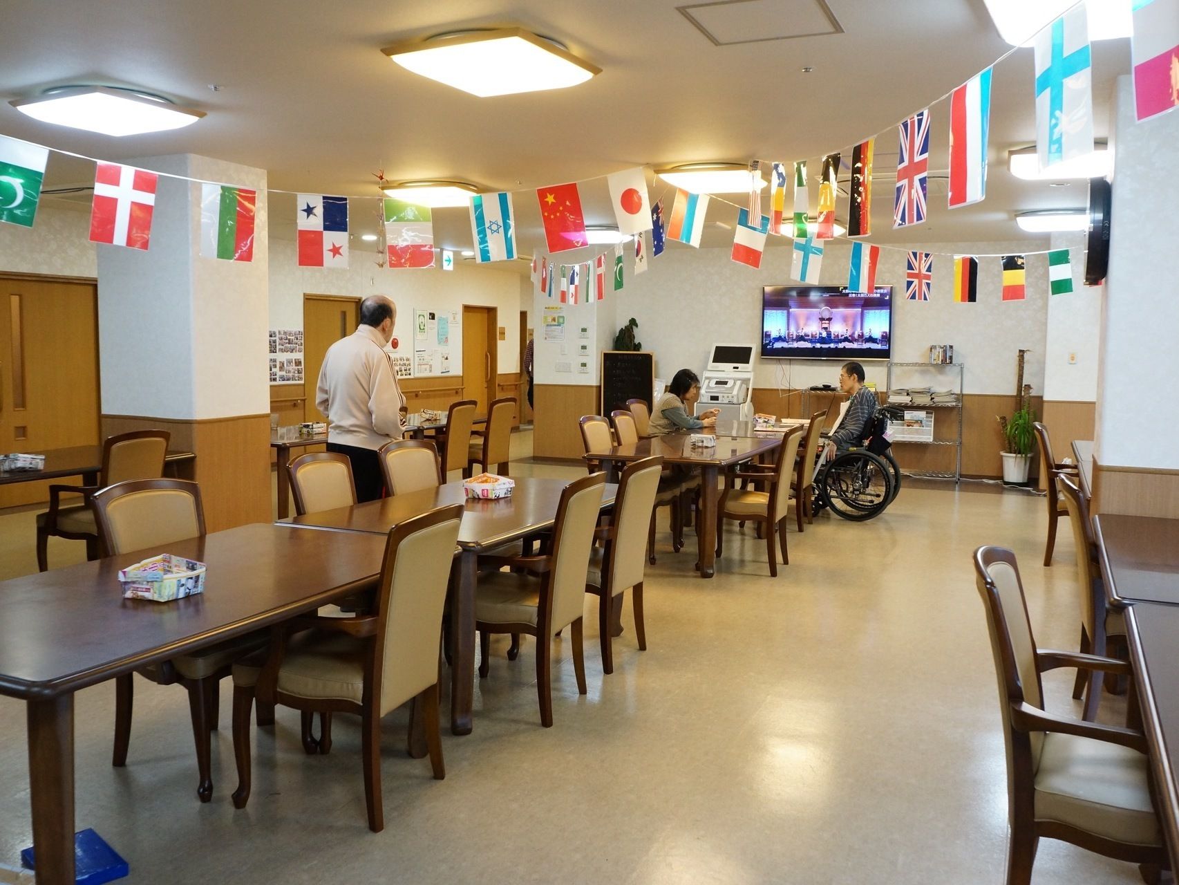 食堂にはテレビを置いており、茶色いテーブルと椅子が並べられている。天井には世界の国旗のガーランドが掛かっている。
