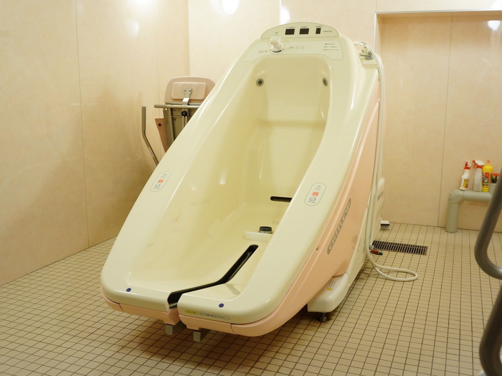グレーのタイルが一面に貼られた浴室の床の中央にはピンク色と白色からなる介護用浴槽が1台置かれている。