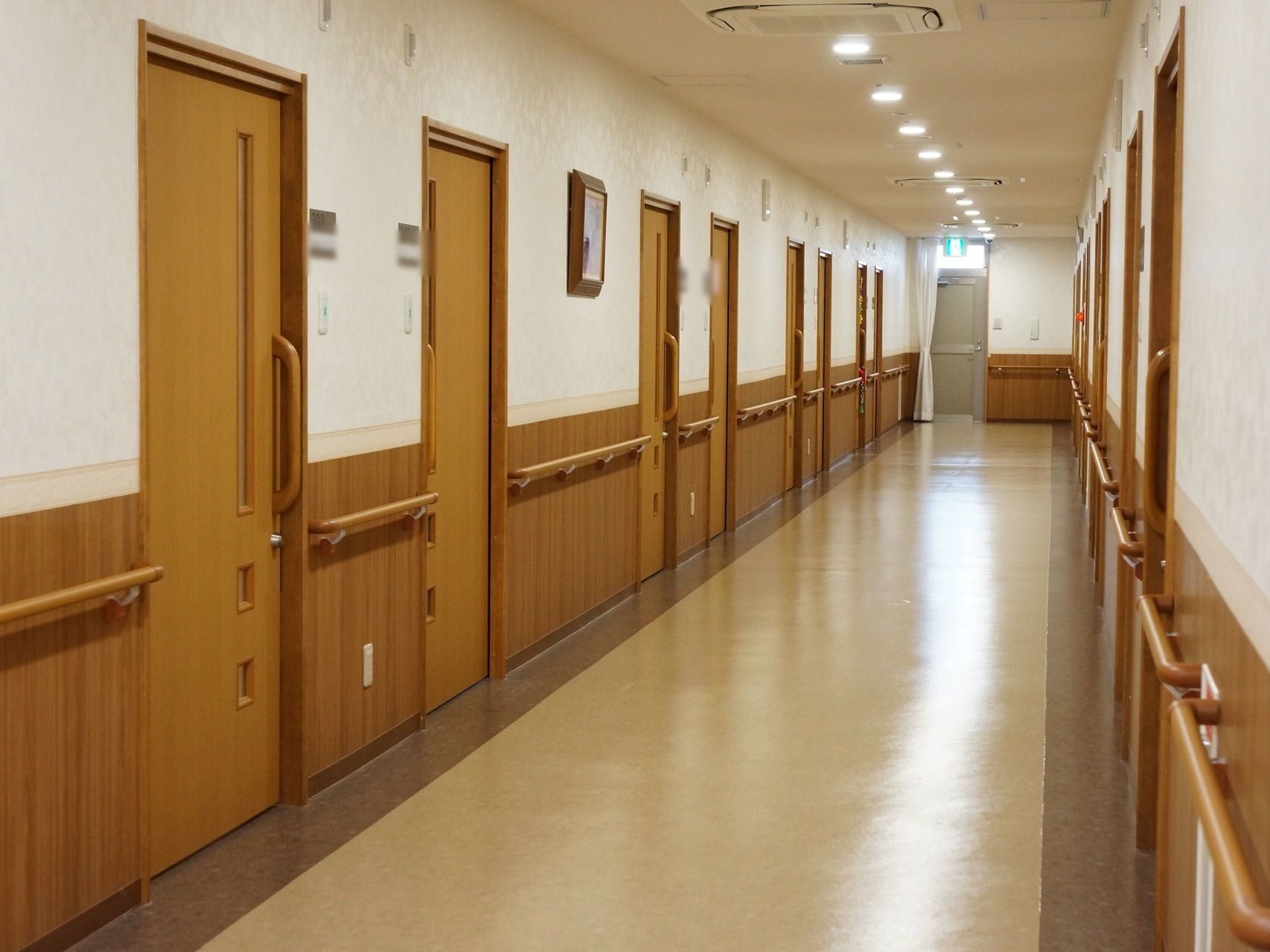 廊下は長く続いている。両側には入居者様の居室がずらりとならんでいる。突き当りには非常口マークのライトが付いている。