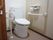 トイレには洋式便器が設置されている。茶色い扉側と反対の壁側には形状がそれぞれ異なる手すりを設置している。
