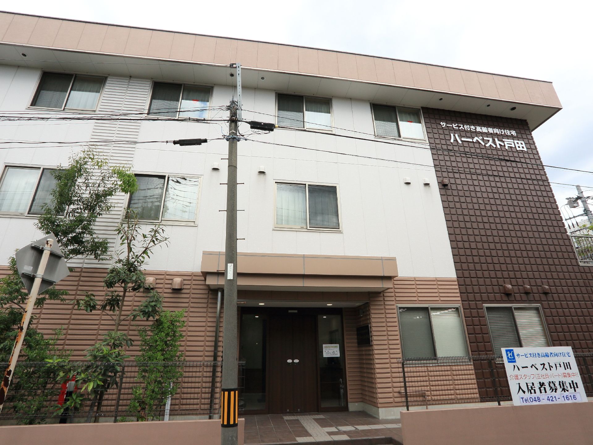 施設は道路に面して建てられている。3階建てで、1階は柵と植え込みで周りをかこっている。入居者募集の看板もある。