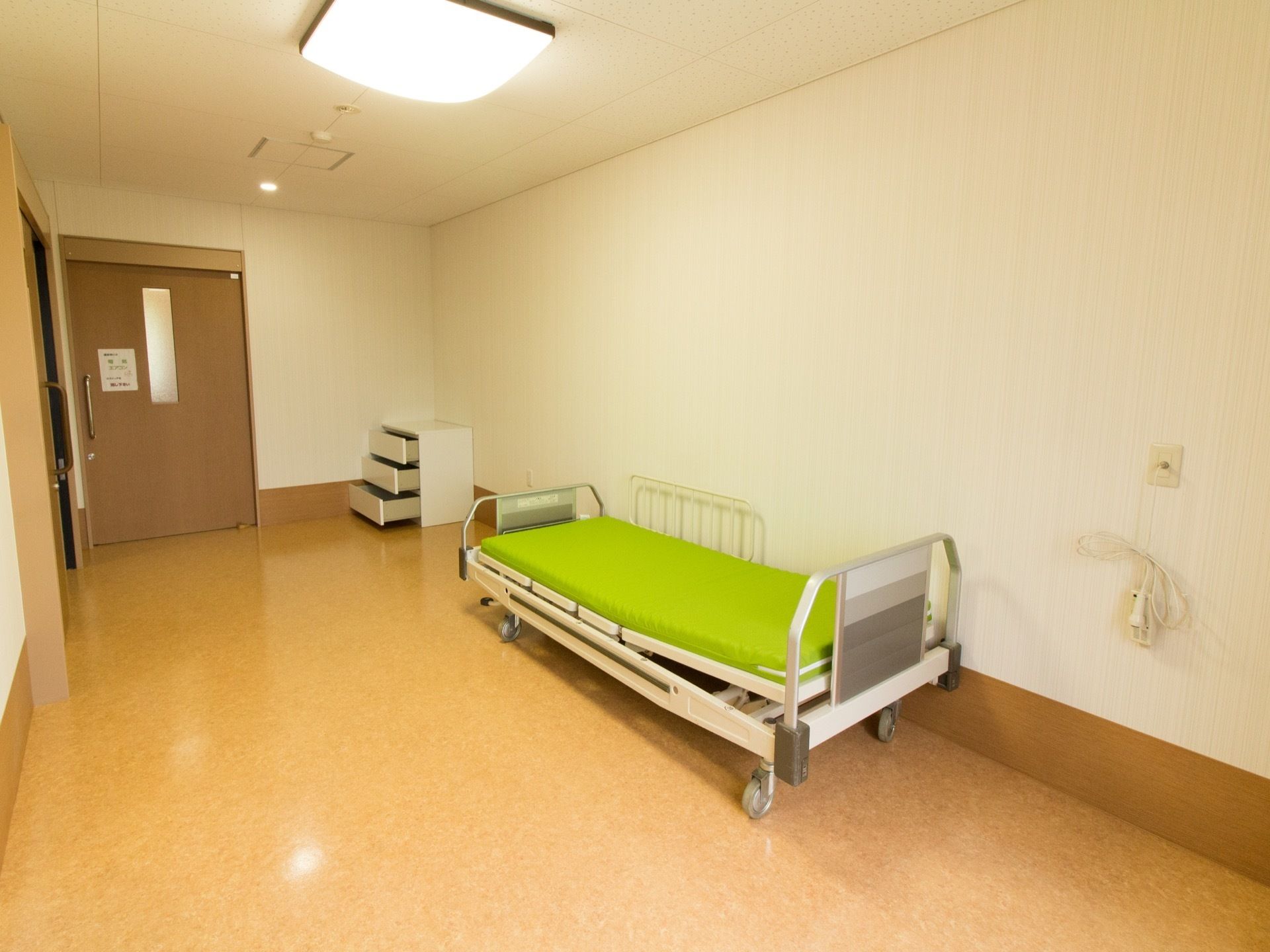介護用ベッドと３段の白いチェストが置かれている。照明は昼光色のシーリングライトの他、入り口付近にダウンライトが設置されている。