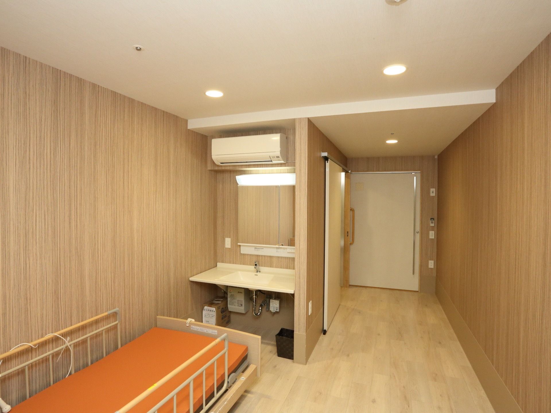 木目を取入れた居室の壁際に介護用ベッドを配置している。横側に洗面台を備え、その上にエアコンを設置している。