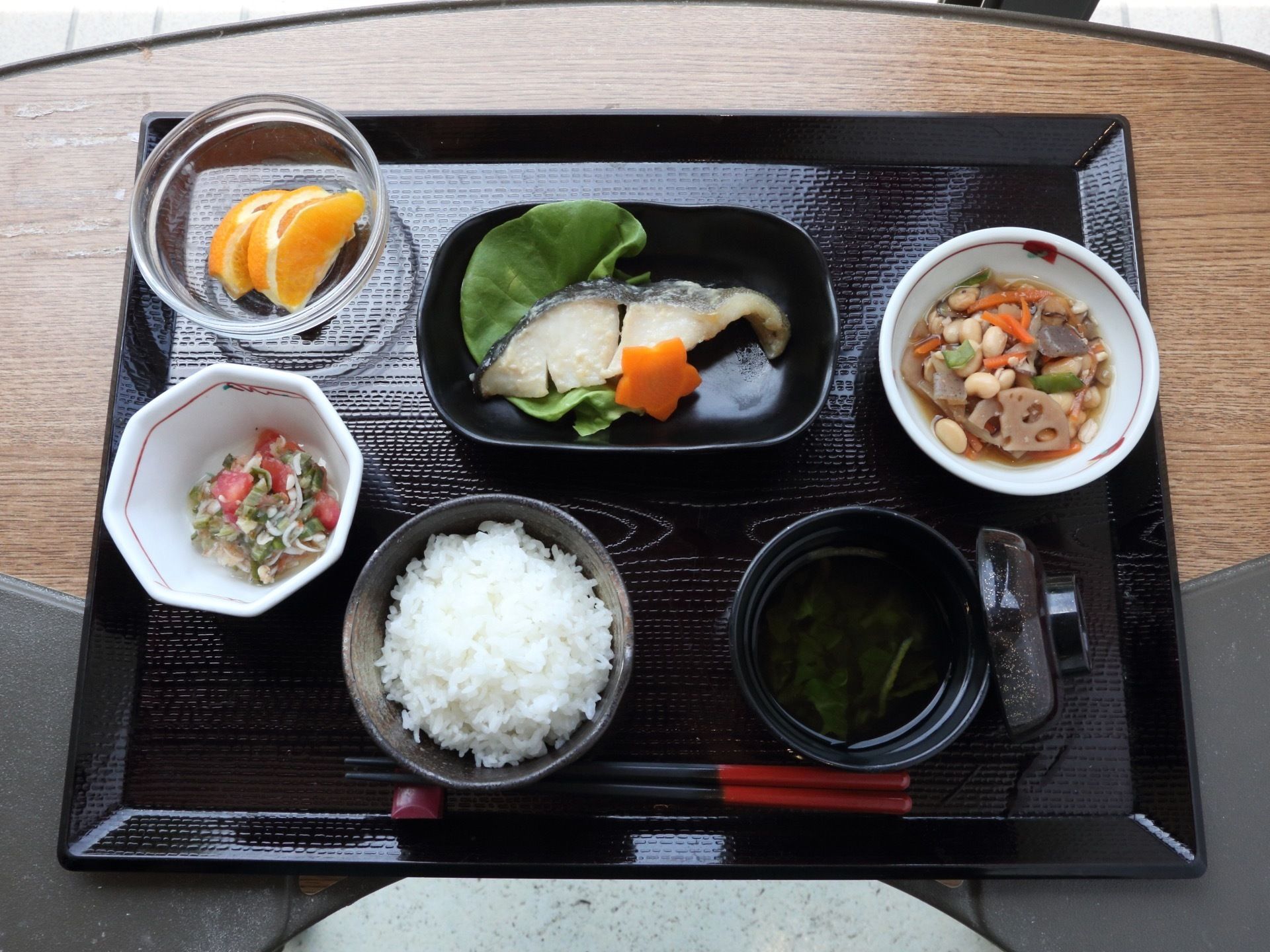 黒のトレーの上にご飯と汁物、焼き魚に和え物、煮物などのおかずとフルーツを添えた食事内容である。彩にも配慮された盛り付けである。