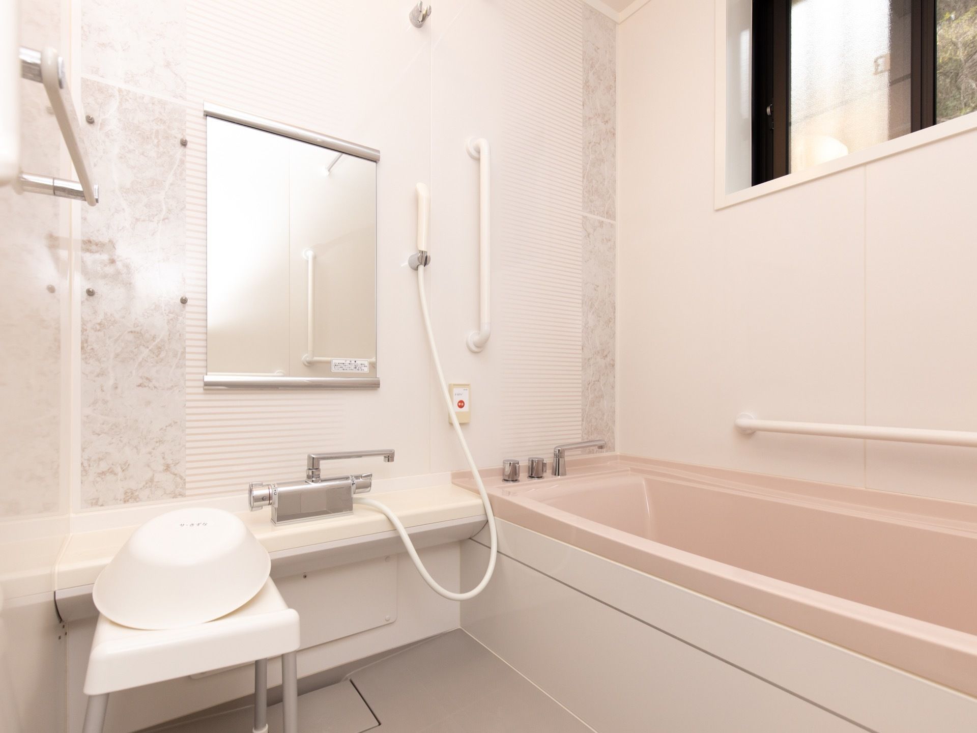 一般家庭にあるような浴室で、洗い場には白い腰かけ椅子と洗面器が用意されている。壁には、緊急通報装置がある。