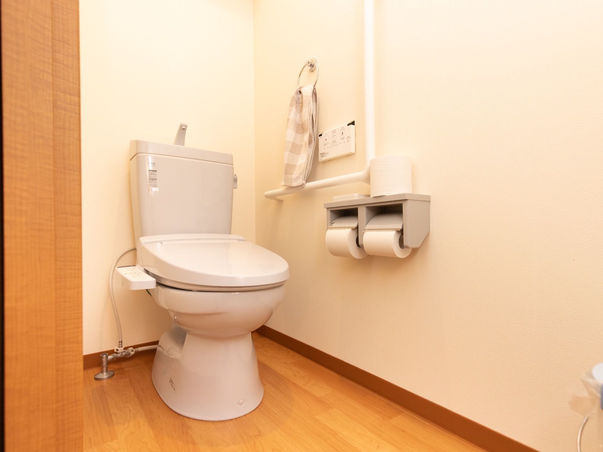 木目のフローリングで、壁は白く塗られている。手すりやシャワー機能の操作盤の近くに、タオルハンガーがある。