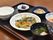 黒いトレイの上にご飯と汁物、真ん中にメインのおかずと２つの小鉢のおかずにお漬物を添えている。手前にお箸を置いている。