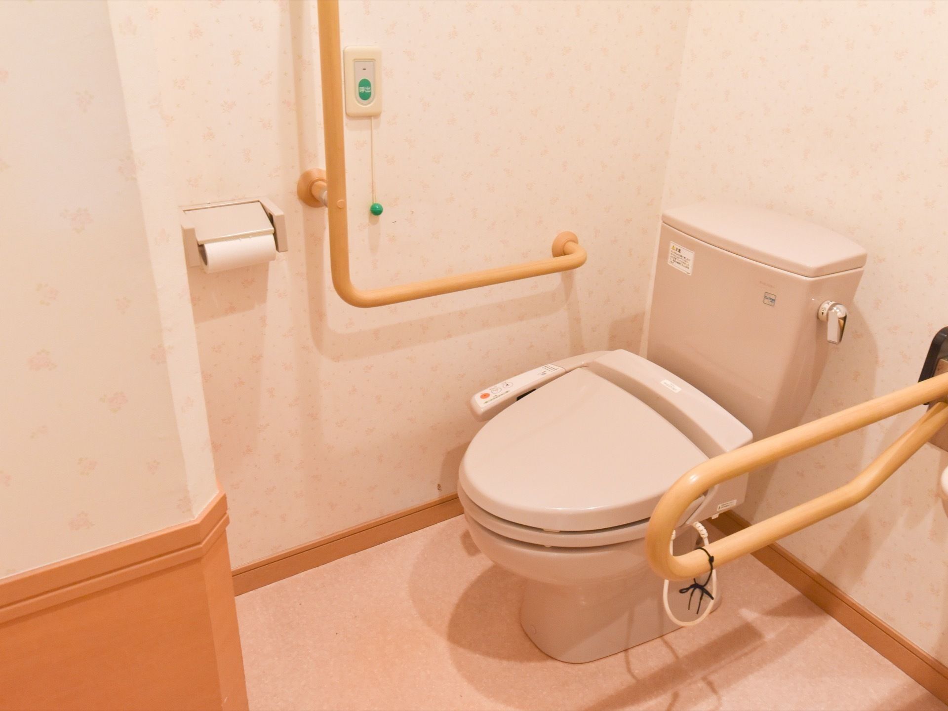 薄いオレンジ色の花柄の壁のトイレである。便器の両側には、茶色の手すりを設けている。壁には、ナースコールを取り付けている。