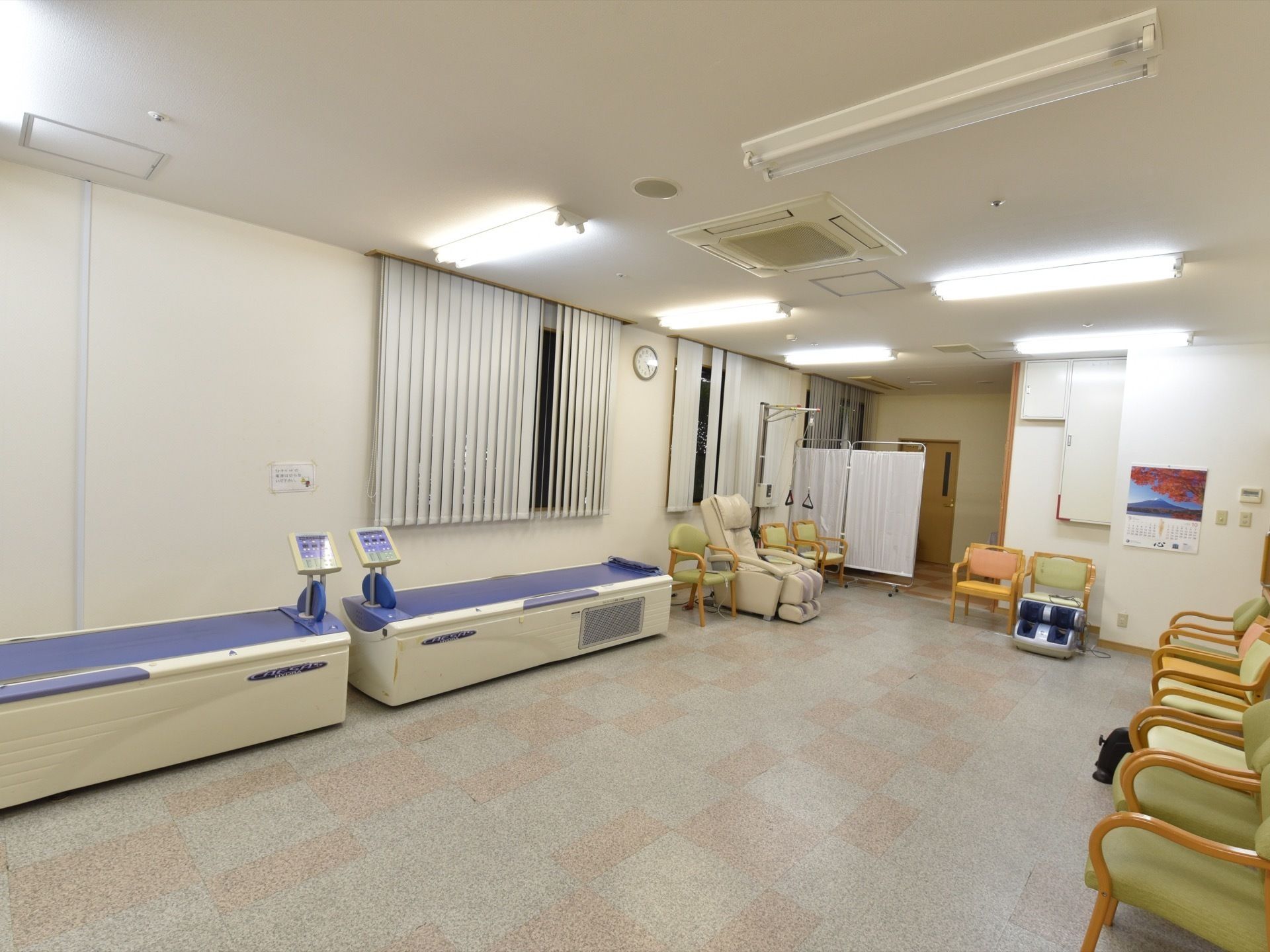 白色の天井と壁にグレイと茶系のデザイン仕様の床のスペースである。中央には広い空間を設けて横に椅子を並べている。機能訓練機器なども置いている。