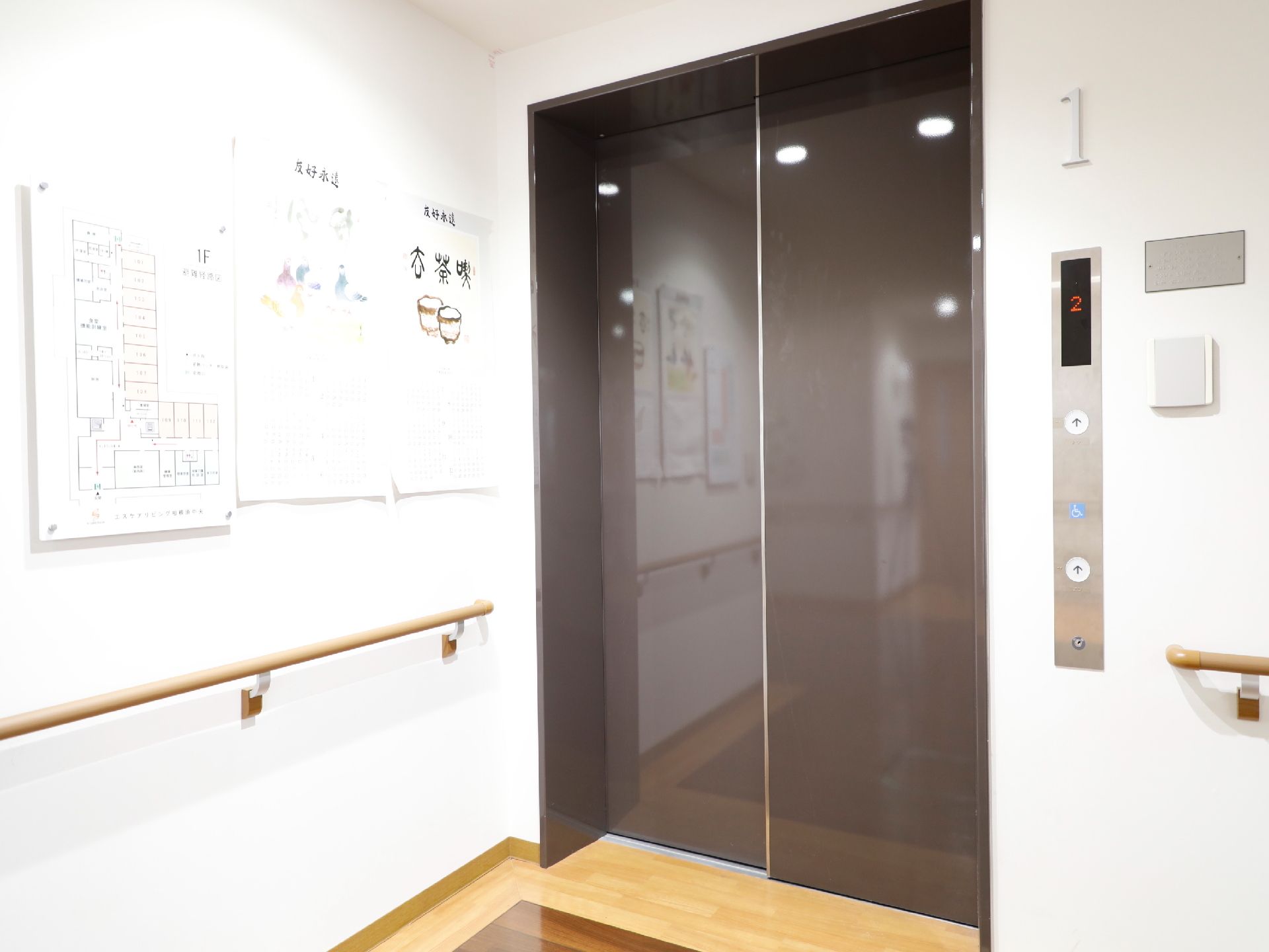 施設には、チョコレート色の扉のエレベーターが設置されている。1階のフロア案内図や転倒防止の手すりが壁に取り付けられている。