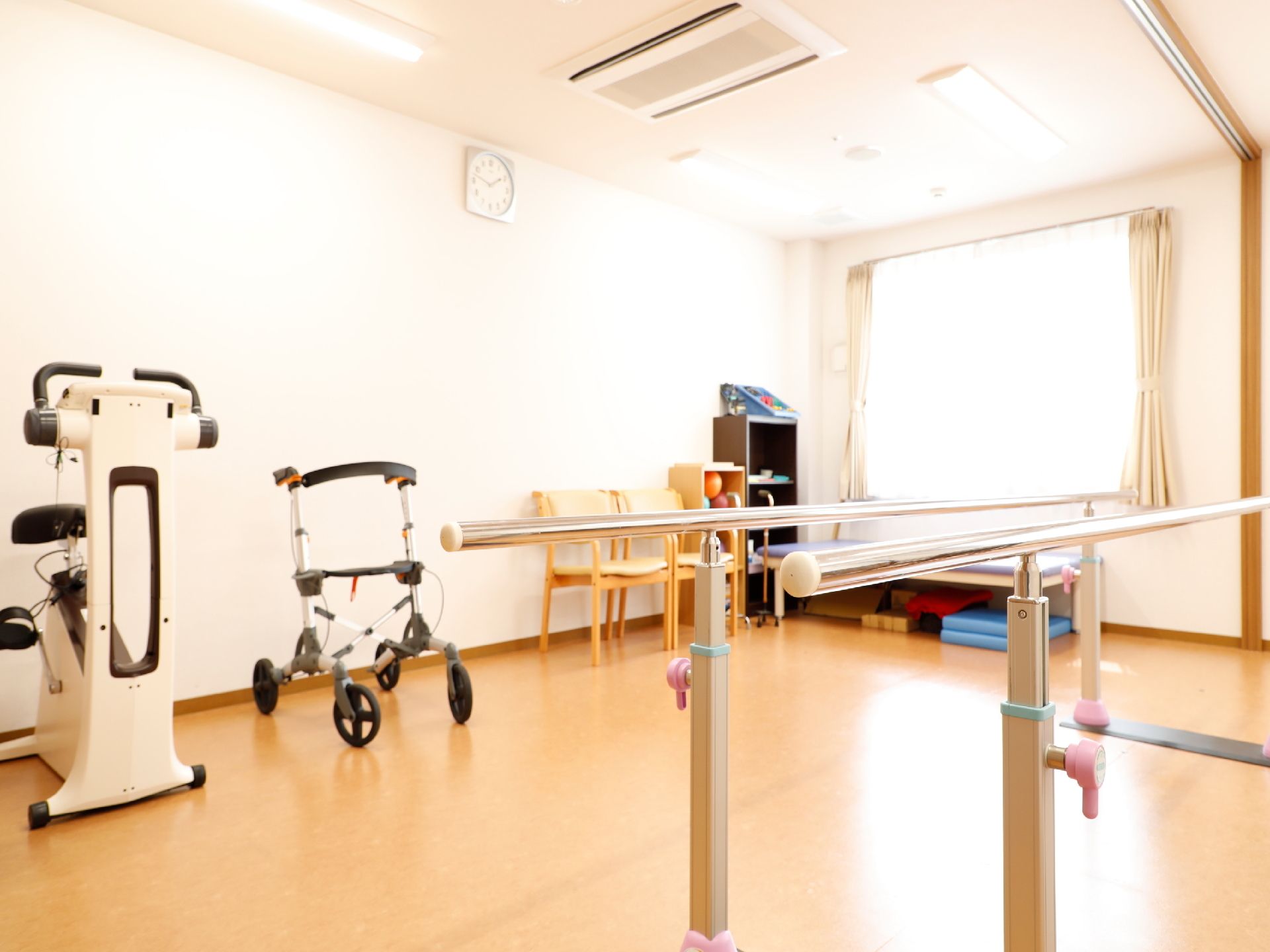 平行棒、歩行器、サイクリング等のリハビリ機器が設置されている。壁沿いにはベッド、四点杖、ボール、一人掛けの椅子が置かれている。