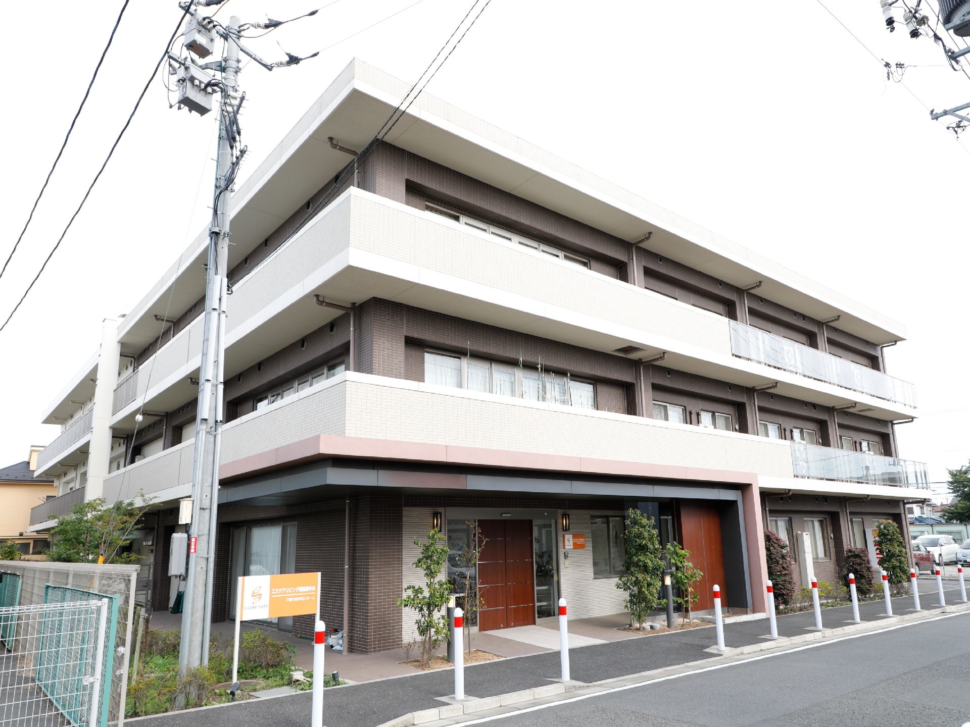 施設は3階建てのしっかりした建物で、目の前には歩道がある。入口付近には、間隔をあけて植物が植えられている。