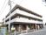 施設は3階建てのしっかりした建物で、目の前には歩道がある。入口付近には、間隔をあけて植物が植えられている。