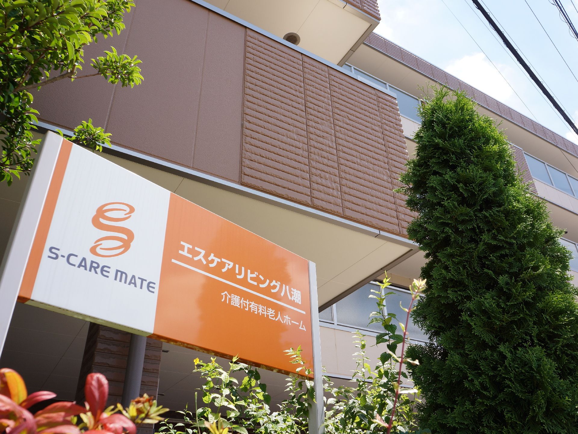 建物前にある施設名が表示された看板の下からのアングルの建物の様子。オレンジ色の看板が、建物の色に合っている。