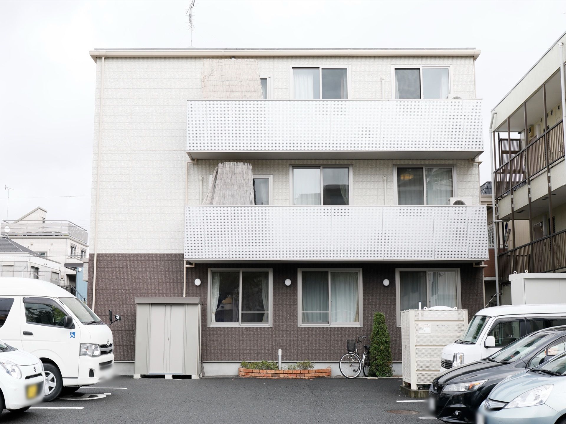 ３階建ての建物には、部屋の窓が均等に並んでいる。窓の外には、駐車場があり向き合ってとまっている車が見えている。