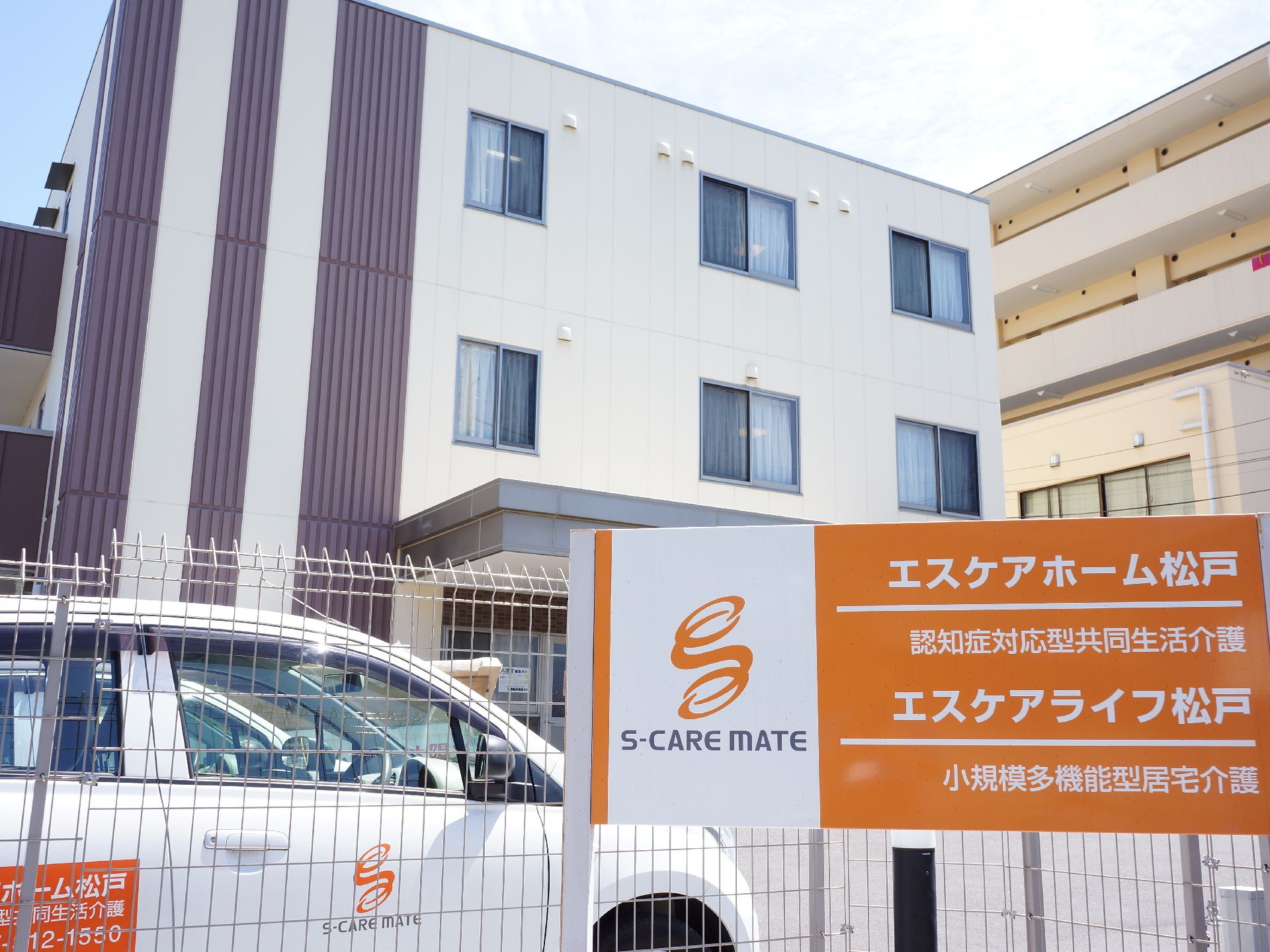 ベージュとダークブランの配色がスタイリッシュな雰囲気の施設の駐車スペースに施設の看板が掲げられている。
