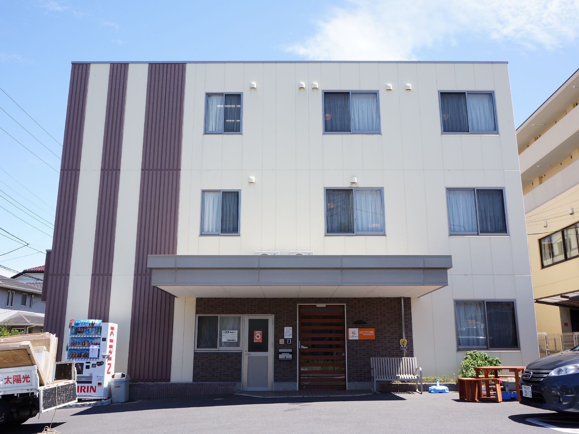 ベージュの建物の外壁にダークブラウンのラインがアクセントになっている建物の正面からの概観。施設の敷地内に駐車スペースが設けられている。