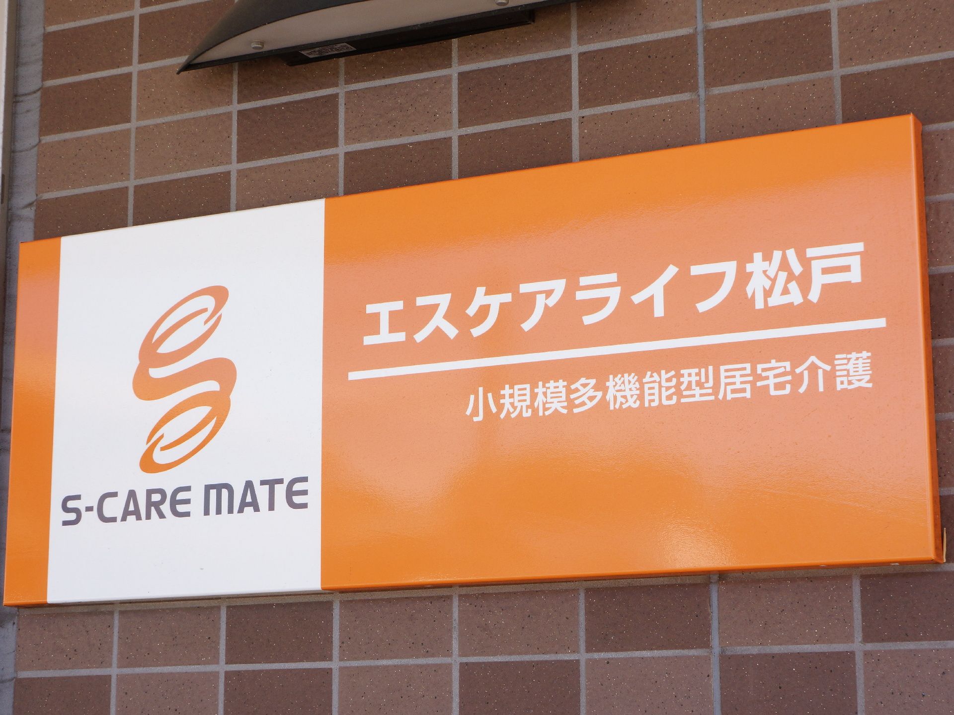 レンガ調の外壁に掲げられた施設の看板。オレンジを基調としたバックにホワイトの文字を使用し明るい雰囲気の看板になっている。