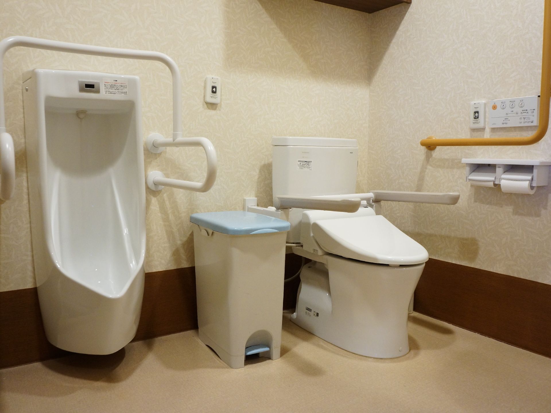 ベージュを基調とした広々としたスペースに男性便器と洋式便器が設置されているトイレ。緊急通報装置が取り付けられている。