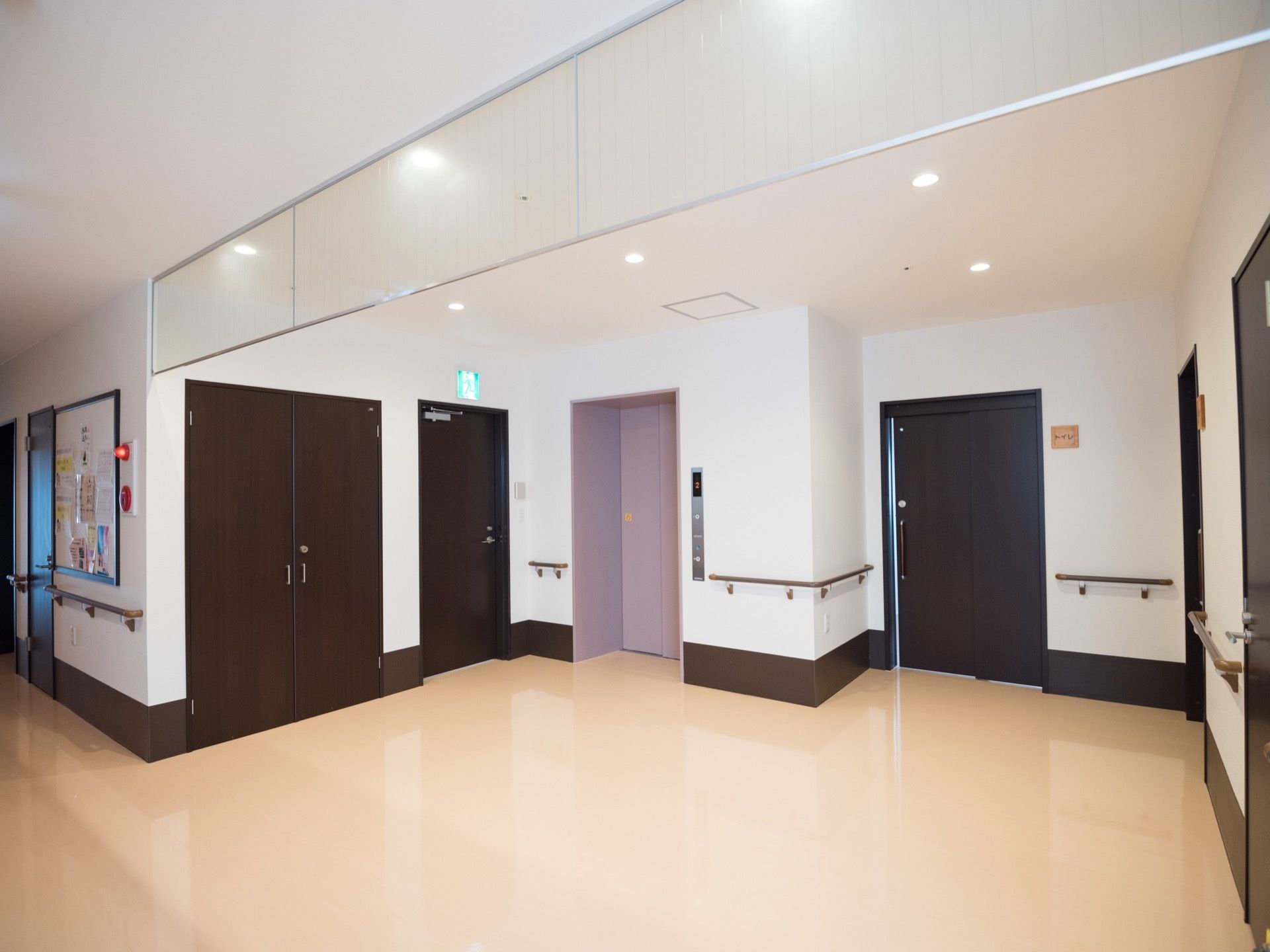 広いエレベーターホールである。横に非常口の標識灯がある。天井に防炎用ガラスが取り付けられている。左側壁には火災報知器が設置されている。
